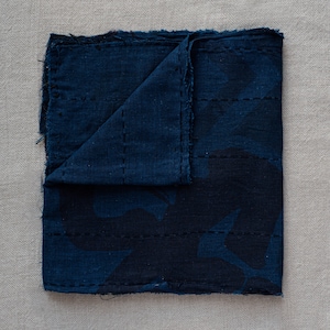 Puede incluir: Una tela de color azul índigo oscuro con un patrón acolchado y bordes deshilachados. La tela está doblada por la mitad y tiene una superficie texturizada.