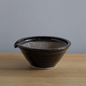 May include: A dark brown ceramic mortar and pestle bowl with a spout. The interior has a textured surface with vertical grooves. The bowl sits on a wooden surface.