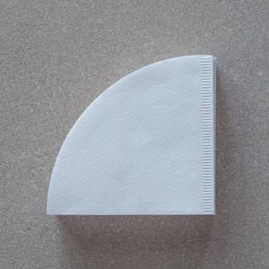 May include: A white paper coffee filter, folded into a cone shape, sits on a grey textured surface.