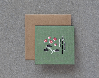 May include: A green card with a black and red illustration of bamboo stalks, clouds, and flowers. The card is surrounded by a brown border.