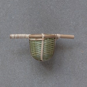 Puede incluir: Un infusor de té de bambú tejido con asa. El infusor es de color marrón claro y tiene un patrón tejido natural.