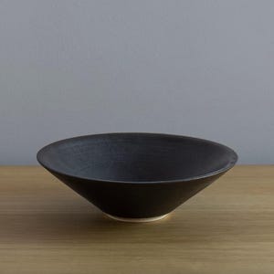 Puede incluir: Un cuenco de cerámica negro con un diseño ancho y poco profundo. El cuenco tiene una superficie texturizada y un borde ligeramente ensanchado. El interior es oscuro y la base es de un color más claro. El cuenco está sobre una superficie de madera.