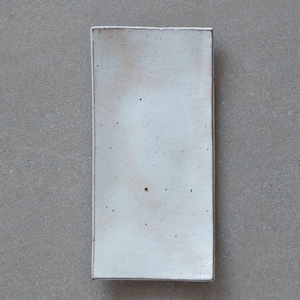 Könnte beinhalten: Rechteckige Servierplatte aus Keramik. Die Platte ist weiß mit einer gesprenkelten Oberfläche und einem dunklen Rand. Das minimalistische Design und die neutrale Farbpalette machen sie vielseitig einsetzbar. Maße: 20 cm x 10 cm.