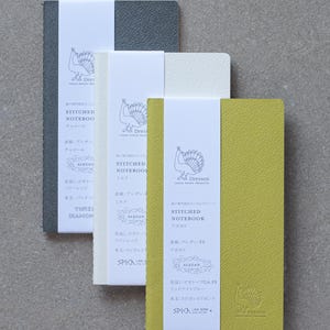 May include: Three stitched notebooks with peacock designs on the covers. The notebooks are in charcoal, white, and avocado green. The covers are textured and the notebooks are labeled "Stitched Notebook" with the color of the notebook listed below. The notebooks are made by Dressco Tafel Paper Products.