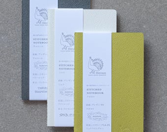 May include: Three stitched notebooks with peacock designs on the covers. The notebooks are in charcoal, white, and avocado green. The covers are textured and the notebooks are labeled "Stitched Notebook" with the color of the notebook listed below. The notebooks are made by Dressco Tafel Paper Products.