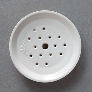 Könnte beinhalten: Eine weiße Keramik-Seifenschale mit mehreren Abflusslöchern. Die Schale hat eine runde Form mit einem leicht erhöhten Rand und einem zentralen Loch. Das Wort "Aloe" ist seitlich eingeprägt.