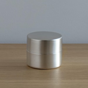 Puede incluir: Un pequeño recipiente cilíndrico de metal cepillado. El recipiente tiene una tapa y se encuentra sobre una superficie de madera. El metal es de color plateado y tiene un acabado mate. El recipiente es de diseño simple y minimalista.