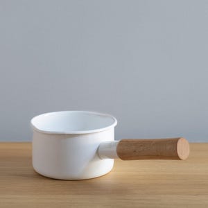 May include: White enamel saucepan with a wooden handle. The saucepan is on a light wood surface.