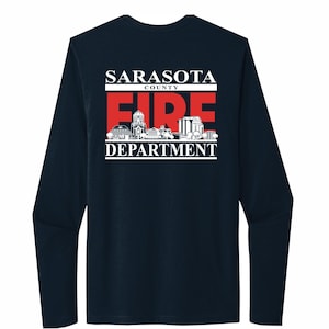 Puede incluir: Camiseta de manga larga azul marino con un gráfico blanco de un horizonte de ciudad y el texto "Sarasota County Fire Department" en rojo y blanco.