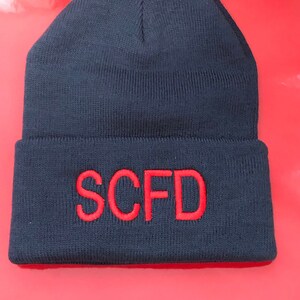 Puede incluir: Gorro de punto negro con un logotipo SCFD bordado en rojo.