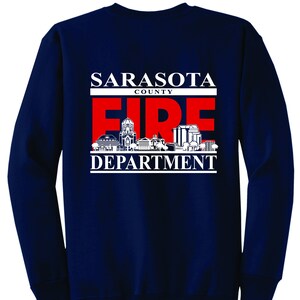 Puede incluir: Sudadera de manga larga azul marino con un gráfico blanco del logotipo del Departamento de Bomberos del Condado de Sarasota. El logotipo presenta las palabras "Sarasota County Fire Department" y un "FIRE" rojo con una silueta de ciudad.