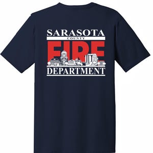 Puede incluir: Camiseta azul marino con un gráfico blanco del logotipo del Departamento de Bomberos del Condado de Sarasota. El logotipo presenta un "FIRE" rojo con una silueta de la ciudad.