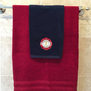 Puede incluir: Una toalla de baño roja con dos rayas blancas colgada de un toallero. Una toalla azul marino más pequeña con un logotipo bordado que dice "Fire Sarasota County Dept." está colgada encima de la toalla roja.