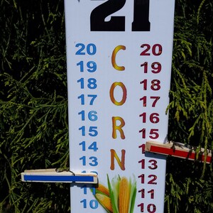 May include: A white vertical cornhole scorekeeper board featuring the number 21 at the top, the word CORN in orange letters, and two columns of numbers from 3 to 20 in blue and red. Two wooden clothespins with blue and red strips mark the scores.
