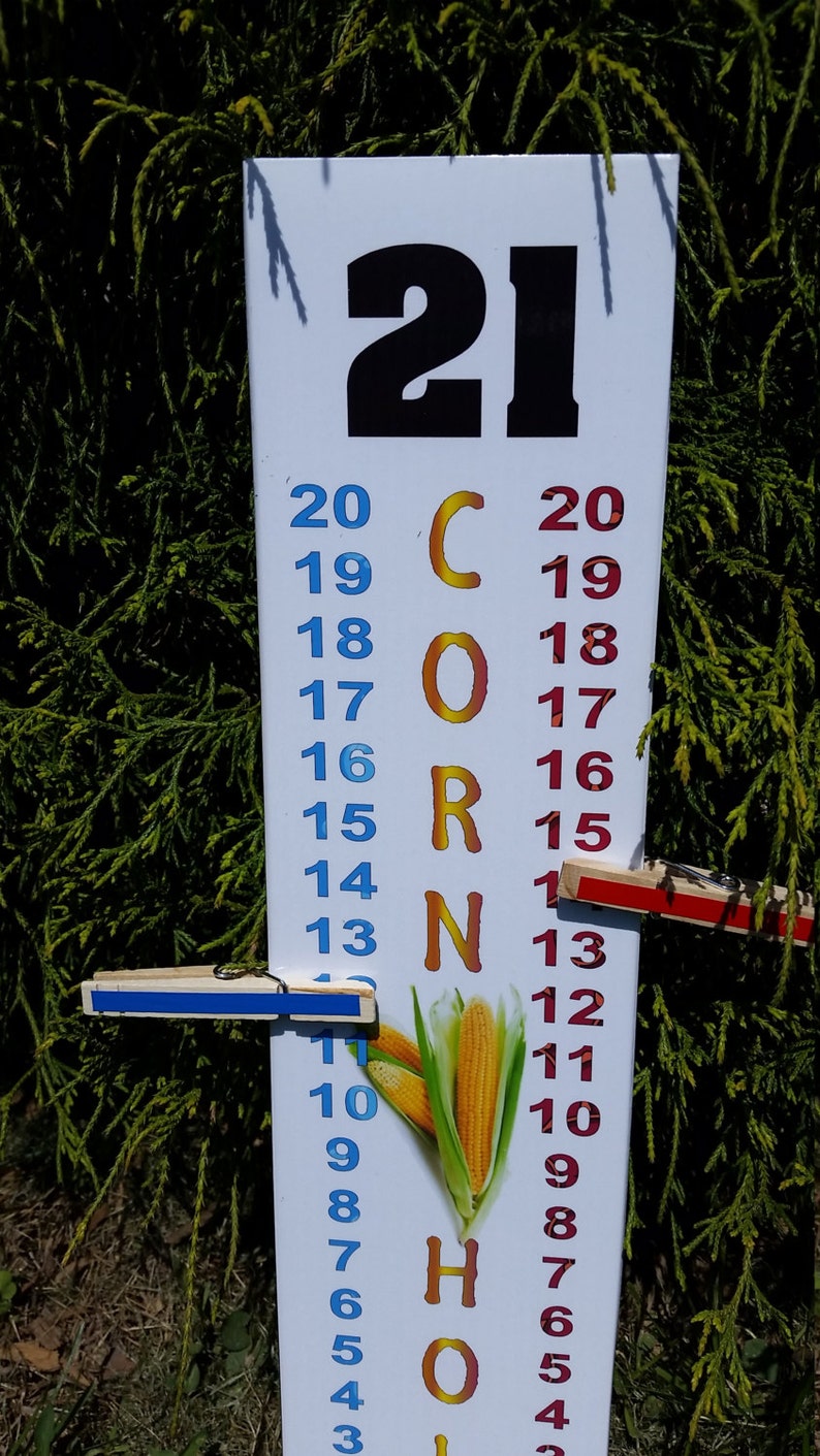 May include: A white vertical cornhole scorekeeper board featuring the number 21 at the top, the word CORN in orange letters, and two columns of numbers from 3 to 20 in blue and red. Two wooden clothespins with blue and red strips mark the scores.