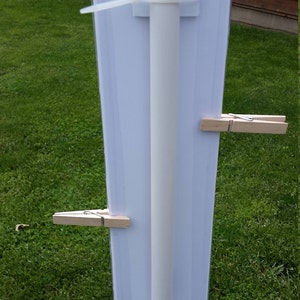 May include: A vertical white rectangular sign attached to a central white pole with two white plastic zip ties. Two light-colored wooden clothespins are clipped onto the sides of the sign, securing a clear plastic sleeve over the white surface.