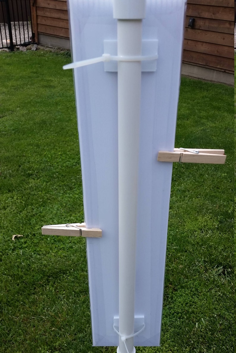 May include: A vertical white rectangular sign attached to a central white pole with two white plastic zip ties. Two light-colored wooden clothespins are clipped onto the sides of the sign, securing a clear plastic sleeve over the white surface.
