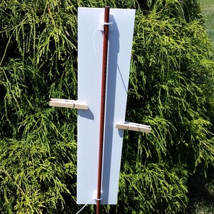May include: A rectangular white sign panel attached to a vertical brown wooden stake. The panel is secured with white plastic zip ties at the top and bottom, and features two light-coloured wooden clothespins clipped to the sides of the board.