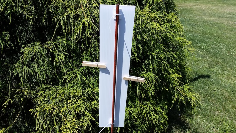 May include: A rectangular white sign panel attached to a vertical brown wooden stake. The panel is secured with white plastic zip ties at the top and bottom, and features two light-coloured wooden clothespins clipped to the sides of the board.