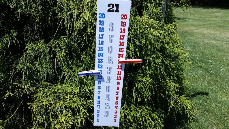 May include: A vertical white scoreboard with the text SCOREBOARD printed in silver. It features two columns of numbers from 1 to 21 in blue and red. A blue clothespin marks the number 12 on the left, and a red clothespin marks 12 on the right.