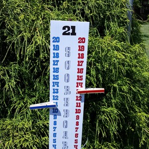 May include: A vertical white scoreboard with the text SCOREBOARD printed in grey. It features two columns of numbers from 0 to 21 in blue and red. Two sliding markers, one blue and one red, are attached to the board to track game scores.