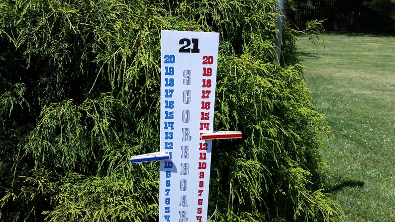 May include: A vertical white scoreboard with the text SCOREBOARD printed in grey. It features two columns of numbers from 0 to 21 in blue and red. Two sliding markers, one blue and one red, are attached to the board to track game scores.