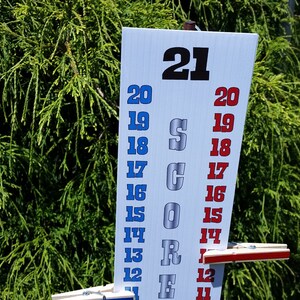 May include: A white vertical score board with the word SCORE printed in large silver letters. The board features two columns of numbers from 11 to 21, with the left column in blue and the right column in red. Two wooden clothespins are attached to the board.