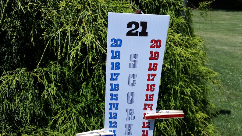 May include: A white vertical score board with the word SCORE printed in large silver letters. The board features two columns of numbers from 11 to 21, with the left column in blue and the right column in red. Two wooden clothespins are attached to the board.