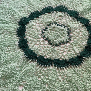 May include: A green and white crocheted doily with a circular design. The doily has a dark green border and a smaller, inner circle.
