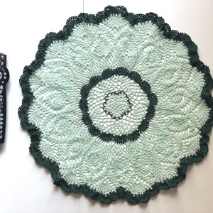 May include: A green and white crocheted doily with a scalloped edge. The doily is round and has a delicate pattern.