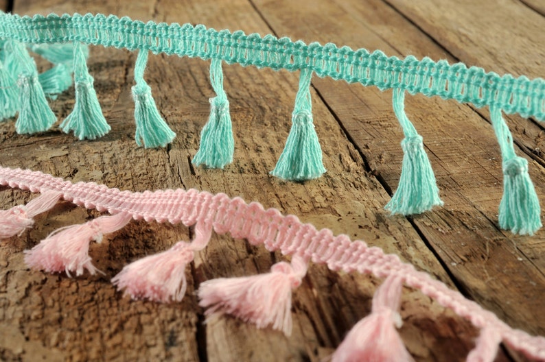 以下が含まれることがあります： 木製の表面に、薄い青とピンクの2種類のフリンジトリムが、タッセル付きで置かれています。