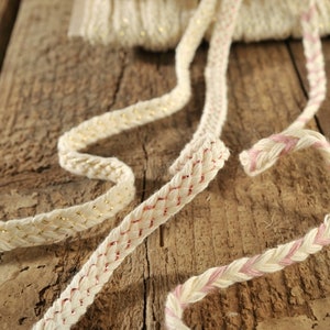 Può includere: Tre tipi diversi di passamaneria intrecciata, ognuno con un accento di colore diverso. La passamaneria è realizzata in fibre naturali e ha un aspetto rustico. La passamaneria è mostrata su una superficie di legno.