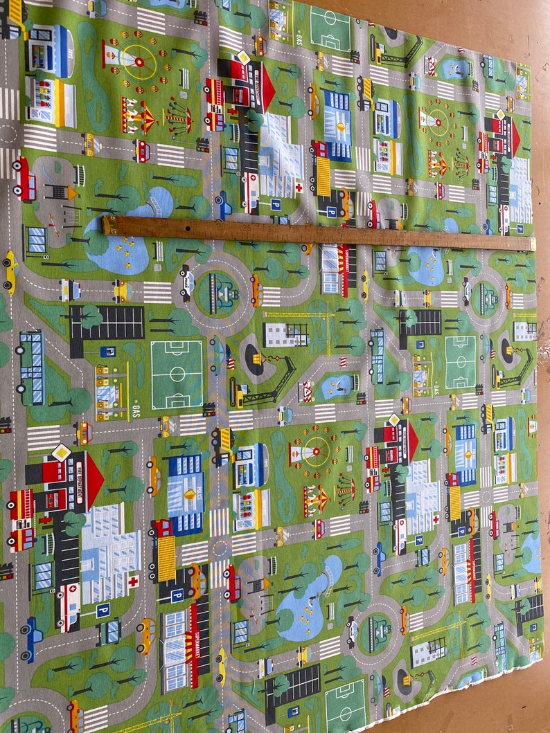 May include: A green and grey playmat with a city scene featuring cars, buildings, trees, and a playground. The playmat has white lines for roads and crosswalks.