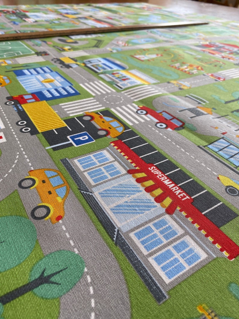 May include: A play mat with a city street design featuring a supermarket, cars, and trees. The supermarket has a red awning with the word "SUPERMARKET" printed on it.