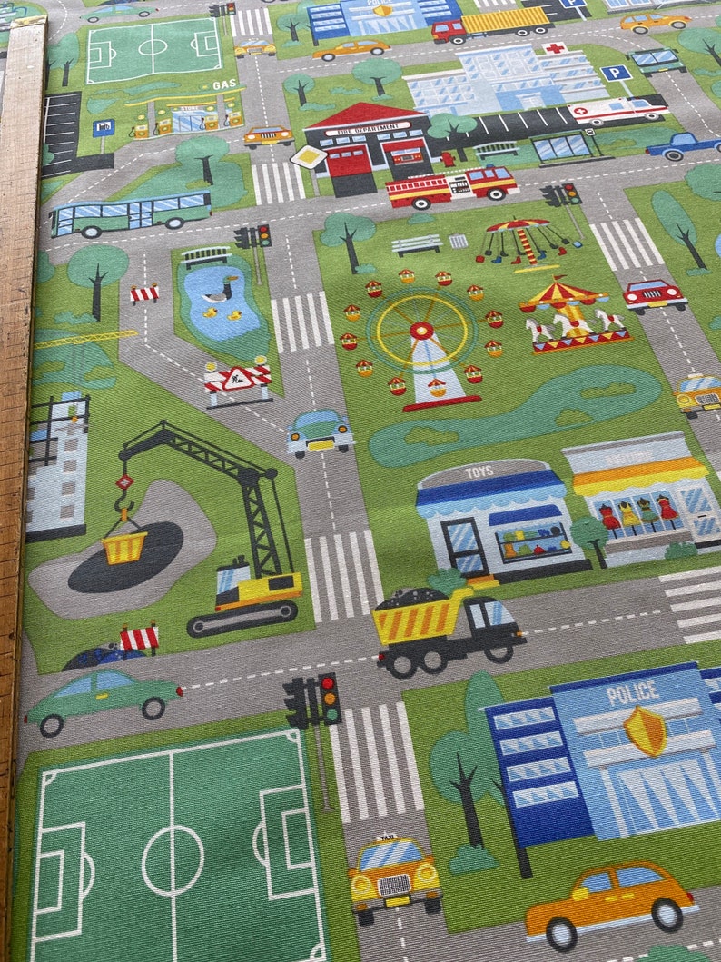 May include: A colourful playmat with a city scene featuring a fire station, a police station, a petrol station, a park, a construction site, and a football pitch. The playmat is made of fabric and has a grey background with white lines.