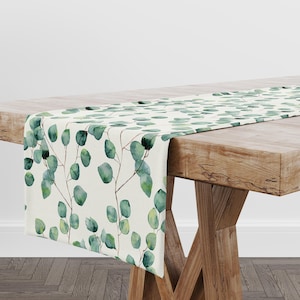 May include: A white table runner with a green eucalyptus leaf pattern. The runner is laid on a wooden table with a natural finish.