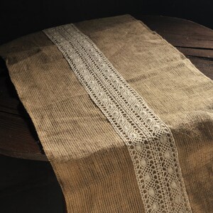 Op de afbeelding: Een bruine jute tafelkleed met een witte kantversiering. De tafelkleed ligt op een donkerhouten tafel.