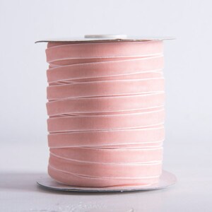 May include: A spool of pale pink velvet ribbon. The ribbon is wound tightly around a white cardboard spool.