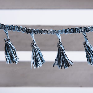 以下が含まれることがあります： 青と黒のタッセルトリムのクローズアップ。複数のタッセルが垂れ下がっています。トリムは織り生地製で、タッセルは柔らかくふわふわした糸でできています。