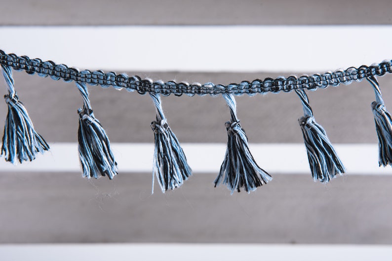 以下が含まれることがあります： 青と黒のタッセルトリムのクローズアップ。複数のタッセルが垂れ下がっています。トリムは織り生地製で、タッセルは柔らかくふわふわした糸でできています。