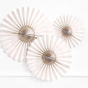 Könnte beinhalten: Drei weiße Papierfächer-Dekorationen, jede in einer anderen Größe, sind an einer weißen Wand angeordnet. Die Fächer bestehen aus gefaltetem Papier und haben ein zartes, geschichtetes Aussehen.