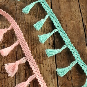 以下が含まれることがあります： 木製の表面に、ピンクとターコイズの2本のタッセル付きトリムが飾られています。トリムは、繰り返しパターンを持つクロシェ編みの生地で作られています。