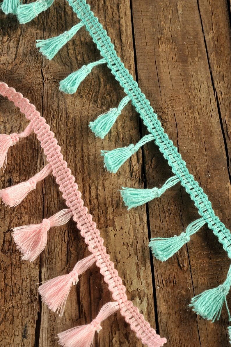 以下が含まれることがあります： 木製の表面に、ピンクとターコイズの2本のタッセル付きトリムが飾られています。トリムは、繰り返しパターンを持つクロシェ編みの生地で作られています。