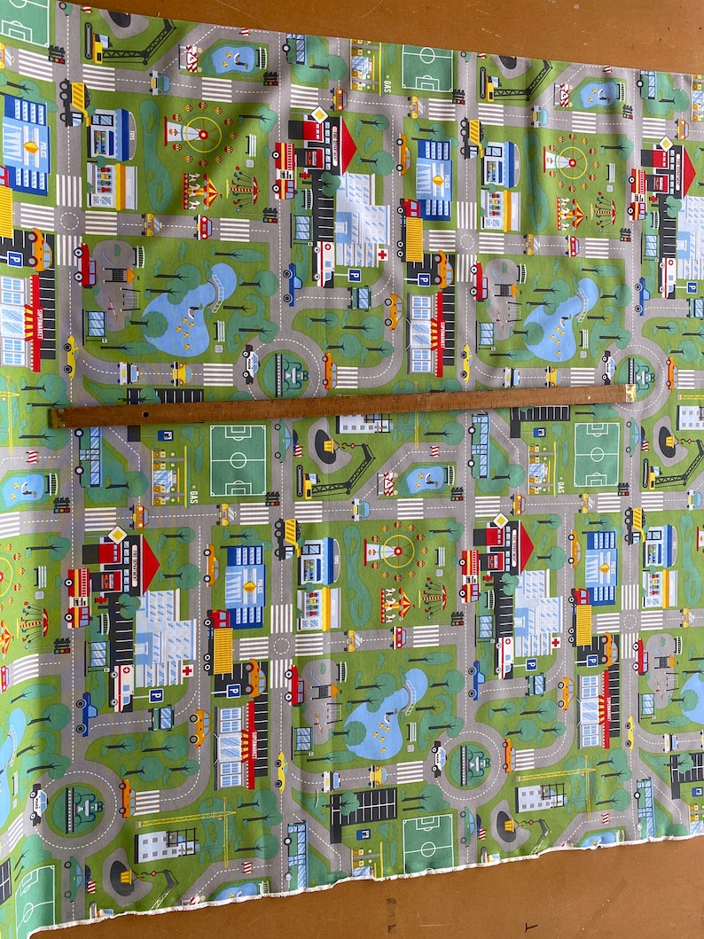 May include: Green fabric with a repeating pattern of a city scene. The pattern includes cars, trucks, buildings, trees, and a playground.