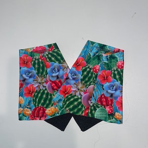 May include: Two fabric pieces with a vibrant cactus and floral print. The design features green cacti, red and blue flowers, and a black fabric detail. The fabric is likely used for crafting or sewing projects.