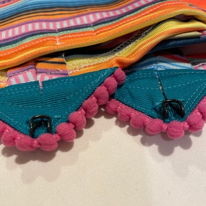K&ouml;nnte beinhalten: Zwei t&uuml;rkisfarbene Stoffkragen mit rosa Bommelbesatz und schwarzen Metallverschl&uuml;ssen. Die Kragen sind &uuml;ber einen bunten gestreiften Stoff gefaltet.
