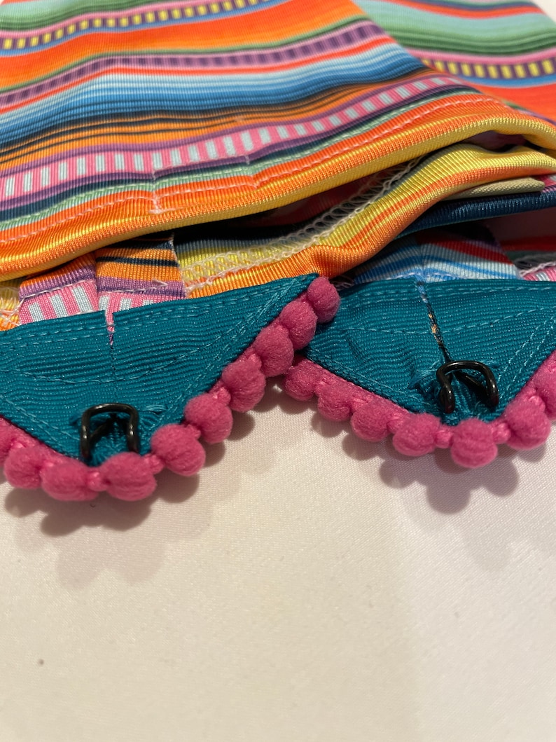 K&ouml;nnte beinhalten: Zwei t&uuml;rkisfarbene Stoffkragen mit rosa Bommelbesatz und schwarzen Metallverschl&uuml;ssen. Die Kragen sind &uuml;ber einen bunten gestreiften Stoff gefaltet.