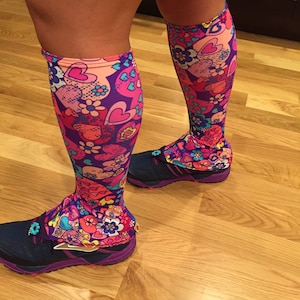 Könnte beinhalten: Ein Paar bunte Beinstulpen mit einem pink-, lila- und blauen Blumenmuster mit Herzen und Blumen. Die Beinstulpen werden über einem Paar dunkelblauer Laufschuhe mit violetten Akzenten getragen.
