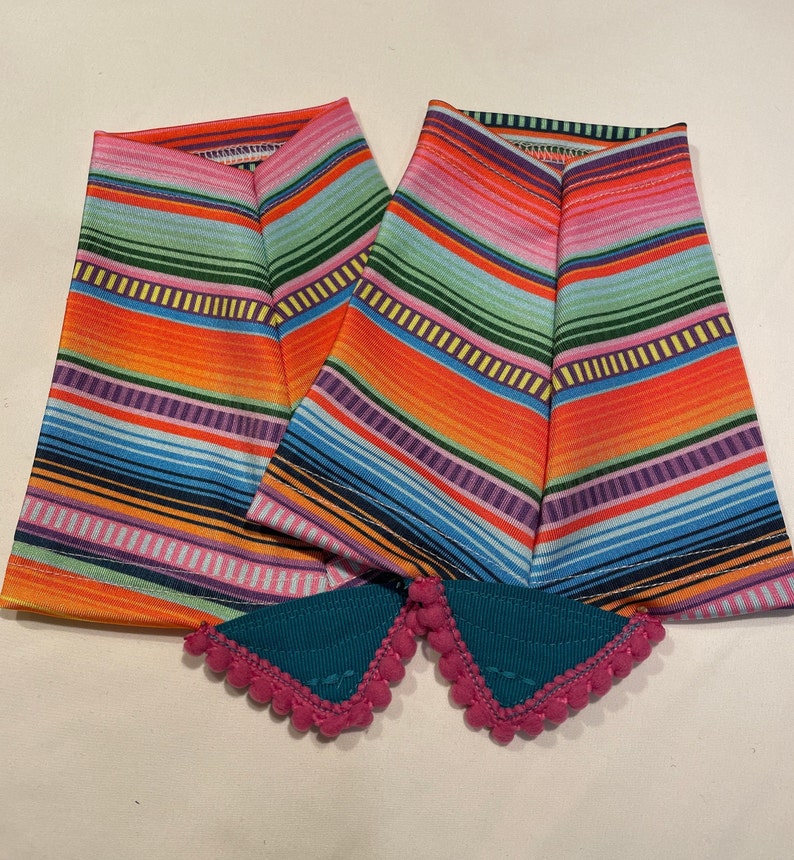 K&ouml;nnte beinhalten: Zwei bunte gestreifte Stoff-Armw&auml;rmer mit rosa Bommelbesatz. Die Armw&auml;rmer haben ein helles orangefarbenes, rosa, gr&uuml;nes, blaues und gelbes Streifenmuster.