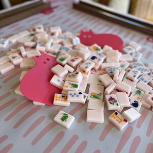 Peut inclure: Un ensemble de tuiles de Mahjong ivoire, avec des symboles et des chiffres color&eacute;s, dispers&eacute;es sur une surface bleu clair avec une d&eacute;coupe rose en forme de chat. Les tuiles pr&eacute;sentent divers motifs, dont des fleurs, du bambou et des points. Un jeu de soci&eacute;t&eacute; classique.